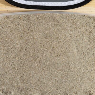 Beiger Spielsand mit feiner Körnung von 0,1-0,8 mm, perfekt für Sandkästen und Spielplätze. Der Sand hat eine sanfte, glatte Textur und ist in einem Behälter zu sehen, der auf einer Holzunterlage platziert ist. Oben im Bild ist der Rand eines Gegenstands mit schwarzem und weißem Stoff zu erkennen. Der natürliche, beige Farbton des Sands erinnert an tropische Strände und ist ideal für kreative Spielprojekte. Verfügbar bei Gartengestaltung Leber in der Steiermark, geeignet für Garten- und Landschaftsbau. leber.at, Steiermark, online kaufen, online bestellen.